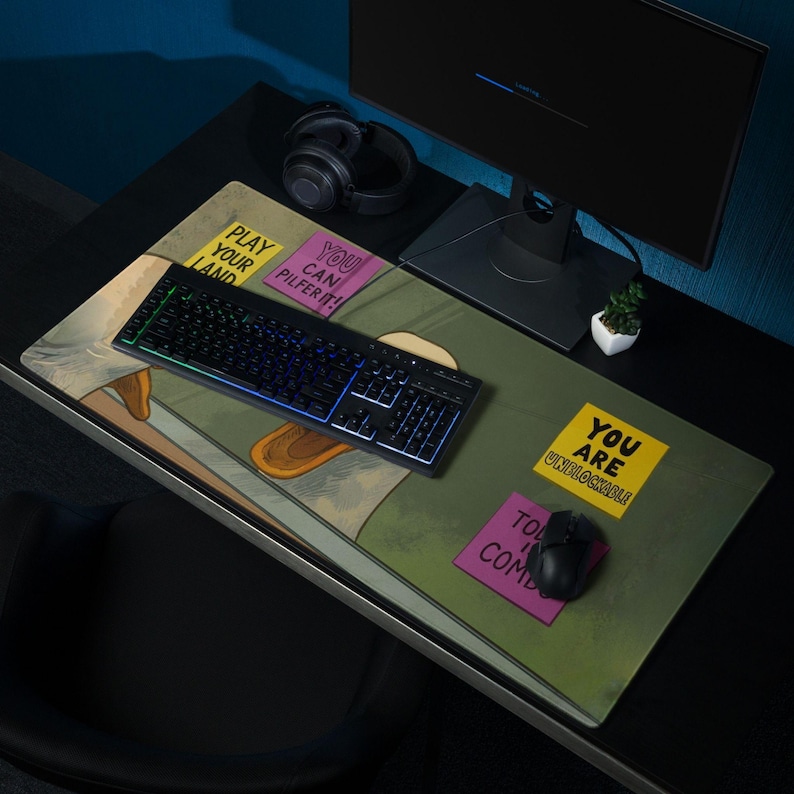 K&ouml;nnte beinhalten: Ein Gaming-Schreibtisch-Setup mit einem gro&szlig;en Mauspad mit Cartoon-Illustration und Text. Eine schwarze Tastatur mit beleuchteten Tasten liegt auf dem Mauspads, zusammen mit einer Maus. Ein Computermonitor und Kopfh&ouml;rer sind ebenfalls sichtbar.