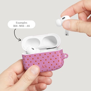 Puede incluir: Una funda rosa para AirPods con un patr&oacute;n repetido de puntos naranjas. La funda est&aacute; abierta, revelando AirPods blancos en el interior. Las iniciales "AS" est&aacute;n impresas en la funda. Una mano sostiene un solo AirPod.