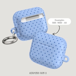 Puede incluir: Dos fundas celestes para AirPods Gen 3 con un dise&ntilde;o de lunares. Una funda tiene un mosquet&oacute;n plateado. El texto "Examples B26-M26-AS" est&aacute; en un &oacute;valo blanco. Las iniciales "AS" est&aacute;n en una funda.