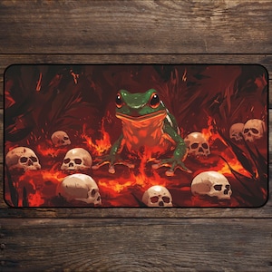 May include: A rectangular mousepad featuring a green frog with red eyes surrounded by skulls and flames. The frog is the central focus, set against a dark red background with a fiery design. The skulls are scattered around the frog.