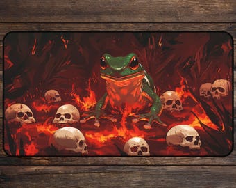 May include: A rectangular mousepad featuring a green frog with red eyes surrounded by skulls and flames. The frog is the central focus, set against a dark red background with a fiery design. The skulls are scattered around the frog.