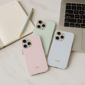 Peut inclure: Trois coques de téléphone pastel avec une texture côtelée. Les coques sont roses, vert menthe et bleu clair, chacune avec un monogramme doré. Un stylo et un carnet ouvert sont également visibles.