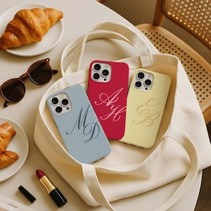 Peut inclure: Trois coques de smartphone en bleu clair, rouge et jaune pâle, chacune arborant un monogramme en lettres cursives. Les coques sont disposées à l'intérieur d'un sac fourre-tout en toile blanche, avec une découpe pour triple objectif photo.