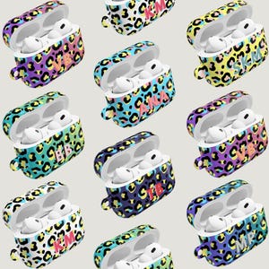 May include: A collection of AirPod cases with a leopard print design in various colors, including blue, yellow, purple, and green. Each case has a different set of initials in a contrasting color. The cases are open, revealing the white AirPods inside.