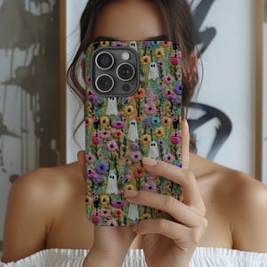 May include: A phone case featuring a colorful floral pattern with small white ghost figures. The case has a variety of flowers in shades of pink, yellow, purple, and blue. The background is a mix of green and brown.
