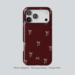 May include: A maroon phone case featuring a repeating design of cream-coloured bows. The case has a camera aperture and the initials "AS" at the bottom. The text at the bottom says "iPhone MagSafe • Samsung Galaxy • Google Pixel".