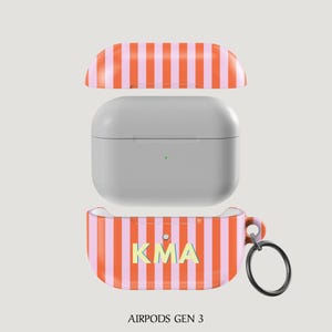 Puede incluir: Funda para AirPods Gen 3 con dise&ntilde;o de rayas naranjas y rosas. La funda est&aacute; abierta, mostrando los AirPods grises en su interior. La parte inferior de la funda tiene las letras "KMA" en amarillo y azul claro. Un llavero negro est&aacute; adjunto.