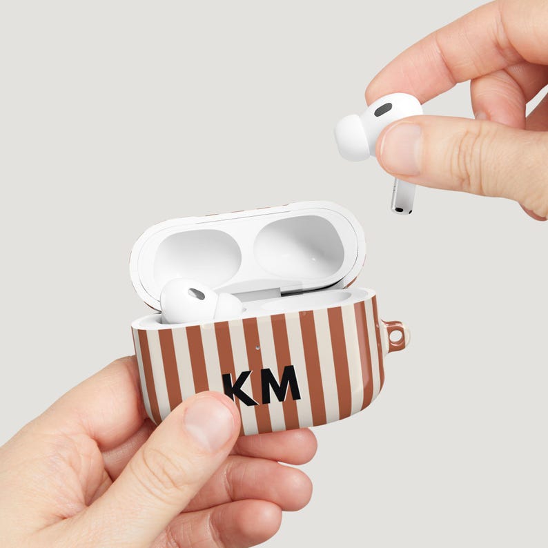 Puede incluir: Auriculares inal&aacute;mbricos AirPods Pro blancos y estuche de carga. El estuche tiene rayas marrones y blancas con las iniciales "KM" en negro. Un AirPod se sostiene en la mano, el otro est&aacute; en el estuche abierto.