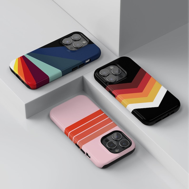 May include: Three phone cases with colorful geometric designs. One case has a rainbow fan pattern, another has a chevron pattern in red, orange, yellow, and white on a black background, and the third is pink with orange stripes.