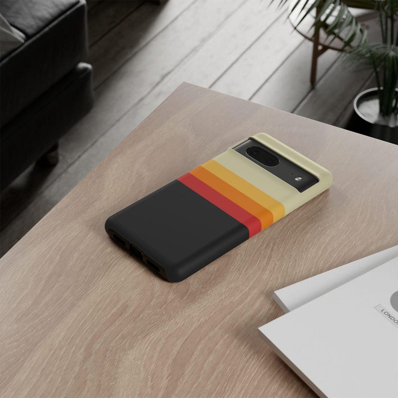 May include: A black smartphone case with a retro stripe design in red, orange, and yellow. The case has a cream-colored top and is resting on a light-colored wooden surface. The case is designed for a smartphone.