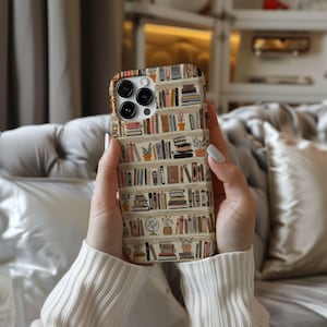 May include: A phone case featuring a bookshelf design with various books and potted plants. The case has a beige background and a colorful array of book spines. The phone case is held in a person's hands.