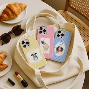 May include: Three phone cases with pet portraits in a white tote bag. The cases are yellow, pink, and blue, each with a different pet portrait and name. A croissant, sunglasses, and lipstick are also visible.