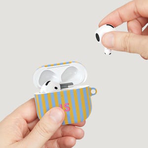 Puede incluir: Una funda para AirPods a rayas azules y amarillas con las letras "LS" en rosa. La funda est&aacute; abierta, revelando AirPods blancos en el interior. Una mano sostiene un solo AirPod. La funda tiene un peque&ntilde;o lazo en el lateral.