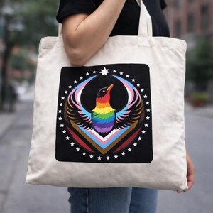 Op de afbeelding: Een crèmekleurige draagtas met een zwarte vierkante print van een vogel met regenboogkleurige veren. De vogel is omcirkeld door witte sterren. De tas wordt door een persoon gedragen, met een stadsstraat op de achtergrond.