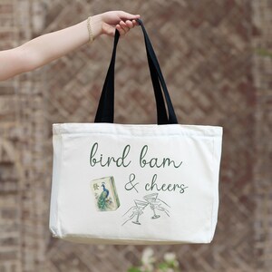 May include: A white canvas tote bag with black handles featuring the dark green cursive text bird bam & cheers. The design includes a small illustration of a mahjong tile with a peacock and a line drawing of two hands clinking martini glasses.