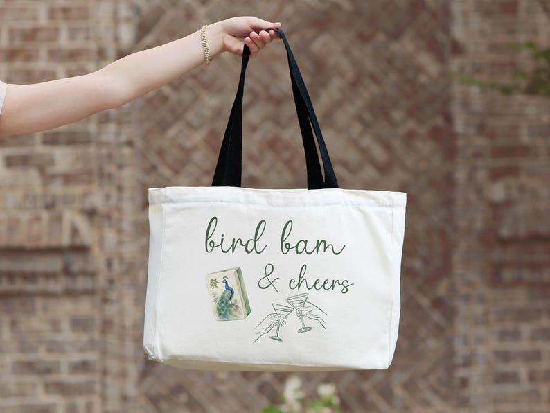May include: A white canvas tote bag with black handles featuring the dark green cursive text bird bam & cheers. The design includes a small illustration of a mahjong tile with a peacock and a line drawing of two hands clinking martini glasses.