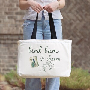 May include: A white canvas tote bag with black handles featuring the green text bird bam & cheers, a small illustration of a peacock mahjong tile, and a line drawing of two hands holding martini glasses in a toast, printed on the front.