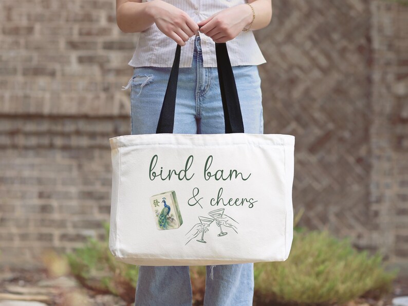 May include: A white canvas tote bag with black handles featuring the green text bird bam & cheers, a small illustration of a peacock mahjong tile, and a line drawing of two hands holding martini glasses in a toast, printed on the front.