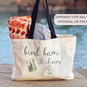 May include: A cream-colored canvas tote bag with black shoulder straps and a zipper closure. The front features the text "bird bam & cheers" in green script, a mahjong tile with a peacock, and two hands holding martini glasses. It has an internal zip pocket.