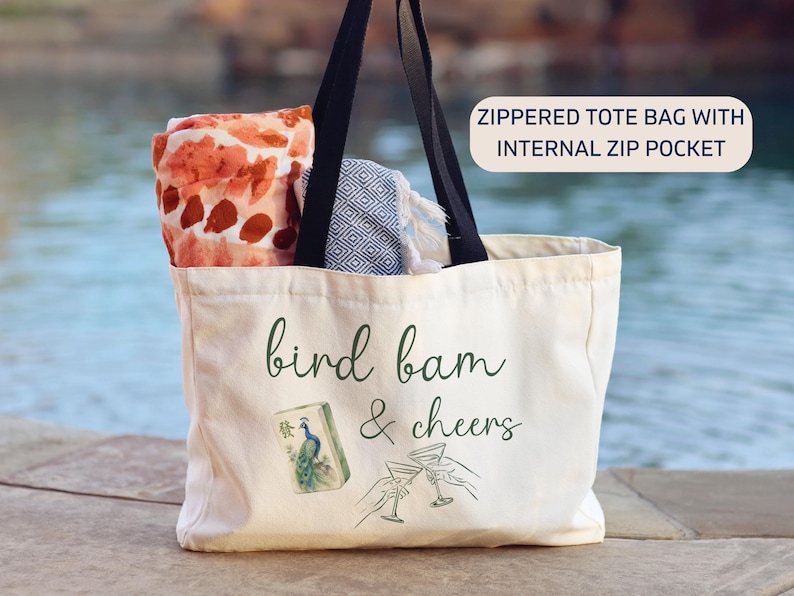 May include: A cream-colored canvas tote bag with black shoulder straps and a zipper closure. The front features the text "bird bam & cheers" in green script, a mahjong tile with a peacock, and two hands holding martini glasses. It has an internal zip pocket.