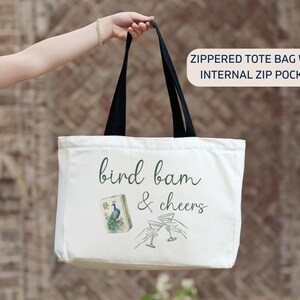 May include: A white canvas tote bag with black handles and a zipper closure. The front features the text "bird bam & cheers" in dark green script, a mahjong tile with a peacock illustration, and a line drawing of two hands clinking martini glasses.