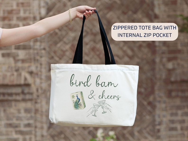 May include: A white canvas tote bag with black handles and a zipper closure. The front features the text "bird bam & cheers" in dark green script, a mahjong tile with a peacock illustration, and a line drawing of two hands clinking martini glasses.