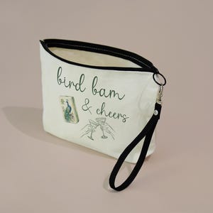 May include: A cream-colored canvas zippered pouch with a black wrist strap. The front features the green text "bird bam & cheers," a small illustration of a mahjong tile with a peacock, and a line drawing of two hands clinking martini glasses.