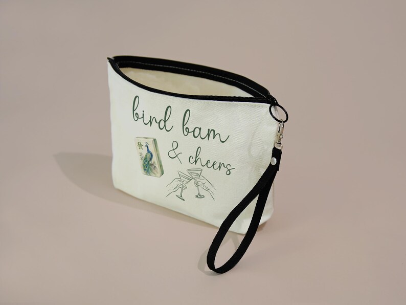 May include: A cream-colored canvas zippered pouch with a black wrist strap. The front features the green text "bird bam & cheers," a small illustration of a mahjong tile with a peacock, and a line drawing of two hands clinking martini glasses.