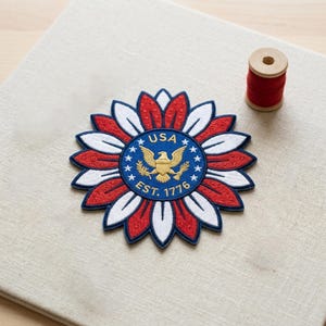 May include: A circular embroidered patch featuring a sunflower design with alternating red and white petals. The center is a blue circle with a gold eagle, the text USA, EST. 1776, and a ring of white stars. A spool of red thread sits nearby.