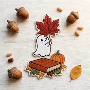 May include: An embroidered patch featuring a white ghost holding a large red maple leaf, positioned above a closed brown book and an orange pumpkin, surrounded by green and yellow autumn leaves, with scattered acorns and seeds nearby.