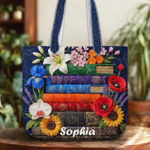 Op de afbeelding: Een marineblauwe draagtas met een ontwerp van gestapelde boeken en bloemversieringen. De tas heeft een witte lelie, blauwe iris, rode klaprozen en zonnebloemen. De naam "Sophia" staat onderaan de tas.