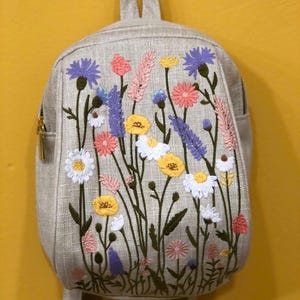 Peut inclure: Un sac à dos beige orné d'une broderie florale colorée. La broderie présente diverses fleurs sauvages dans des tons de rose, jaune, violet et blanc. Le sac à dos est doté d'une poignée supérieure, d'un compartiment principal zippé et de bretelles réglables.