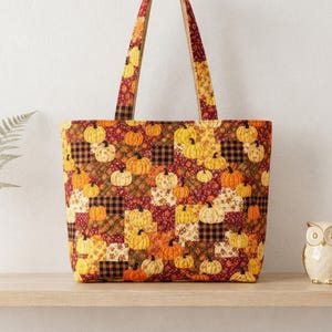 May include: A tote bag featuring a patchwork design with various fabric squares in shades of orange, brown, yellow, and cream. The pattern includes illustrations of pumpkins, black and white buffalo check squares, and floral motifs.
