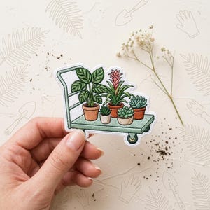 May include: An embroidered patch featuring a light green utility cart carrying five potted plants, including a tall leafy plant, a pink-tipped bromeliad, and three small succulents in various shades of green, all set in terracotta-colored pots.