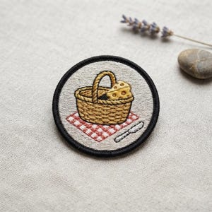 May include: A circular embroidered patch featuring a tan woven picnic basket filled with yellow Swiss cheese, resting on a red and white checkered picnic blanket, accompanied by a small silver knife, all enclosed by a thick black stitched border.