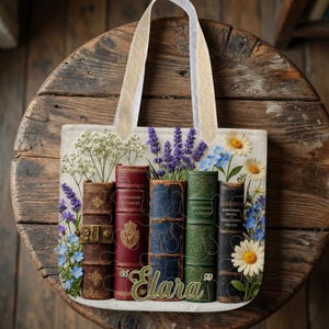 May include: Tote bag with a cream background, depicting a stack of vintage books with floral details like lavender and daisies. The bag has cream handles and the name "Elara" in green script. The design evokes a classic, literary theme.