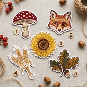 May include: Five embroidered patches featuring a red and white mushroom, a fox face, a yellow sunflower, a bundle of wheat stalks, and an oak leaf with an acorn. Each patch is numbered one through five with small wooden discs on a fabric surface.