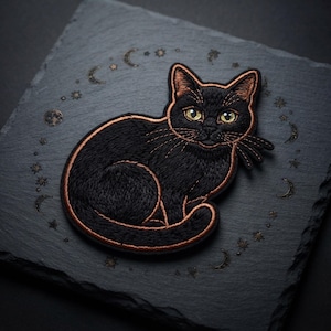 May include: An embroidered patch featuring a black cat with yellow eyes and copper-colored outlines. The cat is depicted in a seated position, surrounded by a circular arrangement of small engraved celestial motifs including moons and stars.