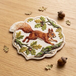 May include: An embroidered patch featuring a detailed ginger squirrel leaping across oak branches with green leaves and small brown acorns. The patch has an off-white background and a scalloped edge, showcasing intricate thread work and textures.
