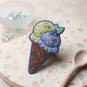 May include: An embroidered patch shaped like an ice cream cone with two scoops. The top scoop is light green with colorful star and sprinkle details, while the bottom scoop is blue with blueberries, stars, and sprinkles. A brown waffle cone base.