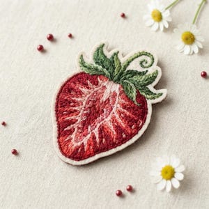 May include: A strawberry patch featuring detailed red and dark pink embroidery of a sliced strawberry with green leaves and a curled stem. The patch has a cream-colored border and is surrounded by small white daisy flowers and red beads.