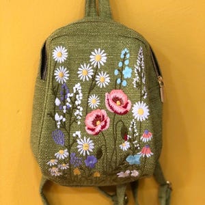 May include: A green backpack featuring embroidered flowers such as daisies and poppies. The bag has a top handle, adjustable straps, and a gold zip. The backpack is displayed against a yellow background.