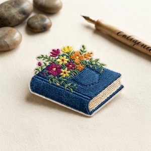May include: An embroidered patch shaped like a blue book with a pocket on the cover, overflowing with colorful yellow, orange, and magenta flowers and green foliage. The book has light tan pages and is positioned near a wooden calligraphy pen.