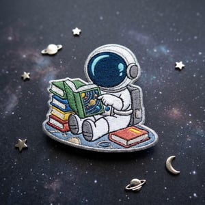 May include: An embroidered patch featuring a white astronaut in a suit with a dark blue visor, sitting and reading a green book with a solar system illustration. A stack of colorful books sits beside the astronaut, with one red book on the ground.