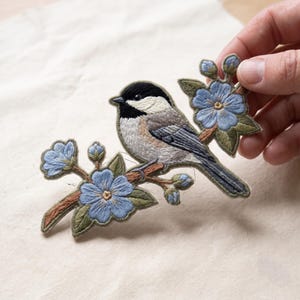 May include: An embroidered patch featuring a black-capped chickadee perched on a brown branch with light blue flowers and green leaves. The bird has a black head, white cheeks, and gray wings. The patch is detailed with intricate thread work.
