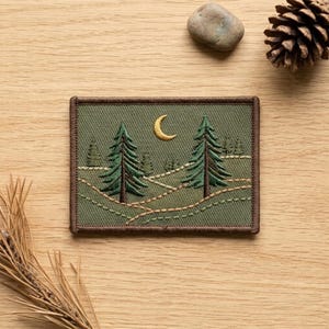 May include: A rectangular embroidered patch featuring an olive green background with two dark green pine trees, a yellow crescent moon, and light green stitched lines representing rolling hills, all enclosed by a dark brown stitched border.