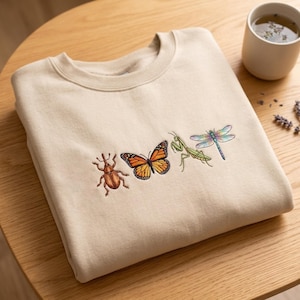 May include: A beige crewneck sweatshirt featuring an embroidered design of four insects arranged in a horizontal row. The embroidery depicts a brown beetle, an orange and black monarch butterfly, a green praying mantis, and a blue dragonfly.