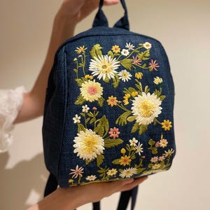 Könnte beinhalten: Ein marineblauer Rucksack mit gesticktem Blumendesign. Die Blumen sind weiß und gelb mit grünen Blättern und rosa und gelben Akzenten. Der Rucksack hat einen oberen Griff und verstellbare Träger.