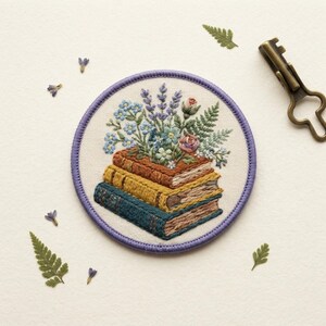 May include: A circular embroidered patch featuring a stack of three books in brown, yellow, and teal, topped with a bouquet of lavender, blue flowers, and green fern leaves. The patch has a purple stitched border and is surrounded by small leaves.