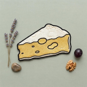 May include: An embroidered patch shaped like a triangular wedge of Swiss cheese with a white top and yellow body featuring dark outlines and holes. The patch is surrounded by a sprig of dried lavender, a smooth brown stone, a walnut, and a grape.